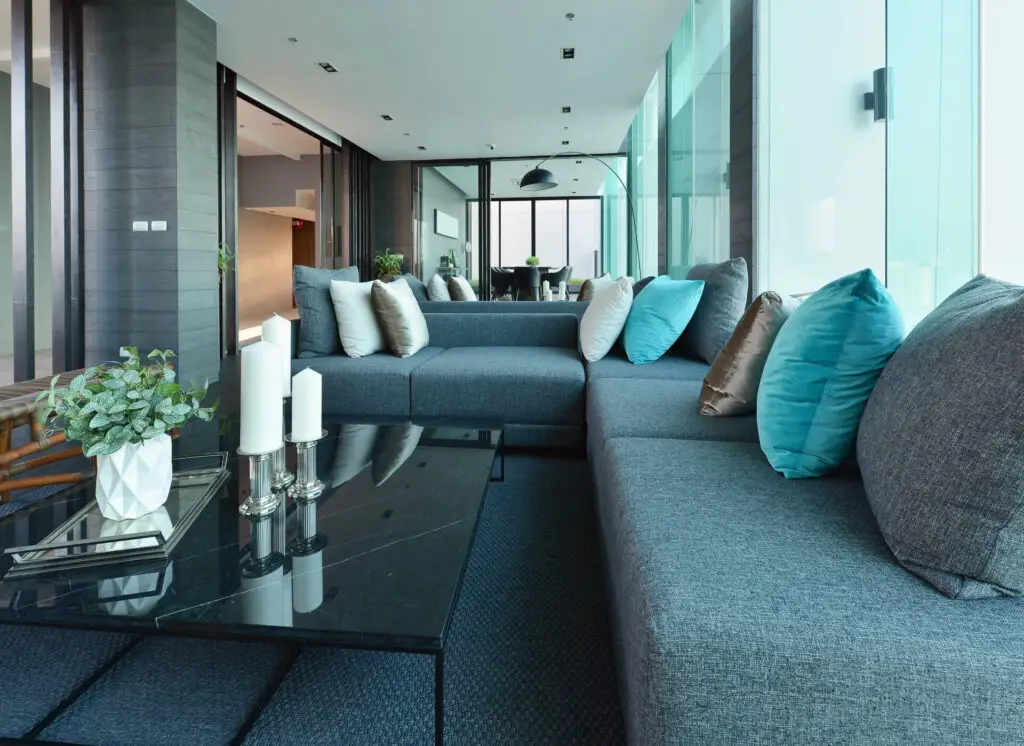 A modern, luxury living room featuring a large grey sectional sofa with teal and white accent pillows, perfect for a real estate agent to showcase to first-time home buyers looking for an upscale, move-in-ready home with floor-to-ceiling windows.