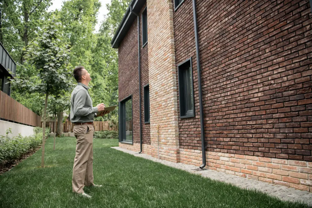 An inspector stands in a grassy backyard holding a clipboard and pen, looking up at the exterior of a two-story modern brick house to determine a home inspection cost