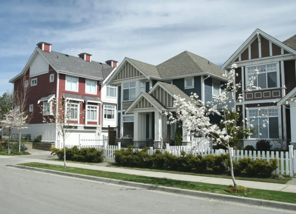 A row of contemporary, multi-story suburban townhomes under a partly cloudy sky, featuring distinct architectural styles and color palettes including deep red, navy blue, and dark brown siding with white trim. The homes are fronted by a neat white picket fence, manicured green hedges, and young trees with white spring blossoms, showcasing a pristine neighborhood setting that reflects the competitive nature of the housing market Tennessee homebuyers are currently navigating.