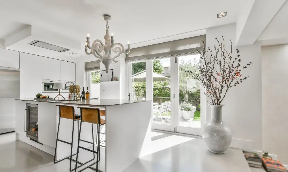 Bright, modern kitchen in a luxury home featuring a white island with bar stools, an elegant chandelier, and large glass doors opening to a scenic backyard patio.